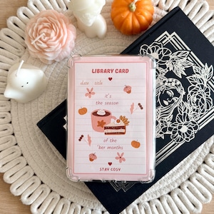May include: A clear phone case featuring a pink library card design with lined paper, autumn illustrations of a mug, cake, pumpkins, and flowers, and the text: LIBRARY CARD, it's the season of the 'ber months, and STAY COSY.