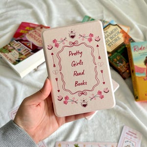 May include: A rectangular, light pink book box with the text "Pretty Girls Read Books" in a decorative frame. The box is surrounded by pink floral and arrow designs. Several books and bookmarks are visible.