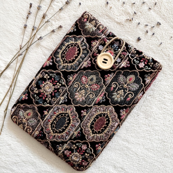 Könnte beinhalten: Eine rechteckige schwarze Stofftasche mit einem komplizierten, sich wiederholenden Blumen- und Geometriemuster in Rosa-, Blau- und Goldtönen. Die Tasche wird mit einer beigen elastischen Schlaufe und einem runden, hellen Holzknopf verschlossen.