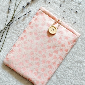 Könnte beinhalten: Eine rosa Stofftasche mit Blumenmuster und einem Verschluss aus Holz. Die Tasche liegt auf einer weißen, flauschigen Oberfläche.