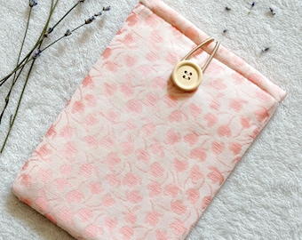 Puede incluir: Una bolsa de tela rosa con un patrón floral y un cierre de botón de madera. La bolsa está sobre una superficie blanca y esponjosa.