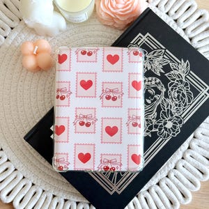 May include: A clear phone case with a pink and white background pattern. The pattern features red hearts, cherries, and bows.