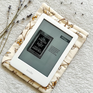 May include: A white Kindle e-reader with a floral patterned case. The screen displays the cover of the book "Through the Mist" by Lindsay Jayne Ashford. The text on the cover reads "A young newlywed discovers that her husband's secrets threaten more than her marriage."