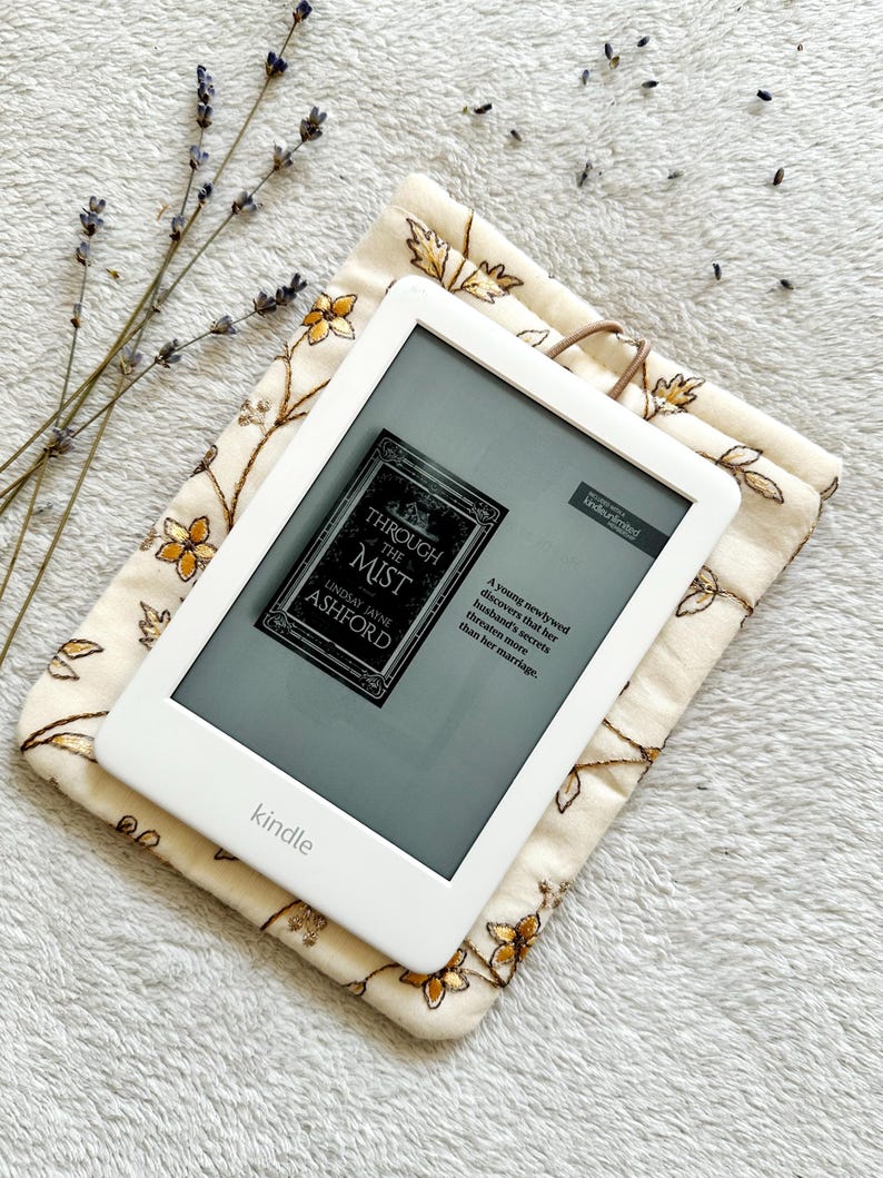 May include: A white Kindle e-reader with a floral patterned case. The screen displays the cover of the book "Through the Mist" by Lindsay Jayne Ashford. The text on the cover reads "A young newlywed discovers that her husband's secrets threaten more than her marriage."