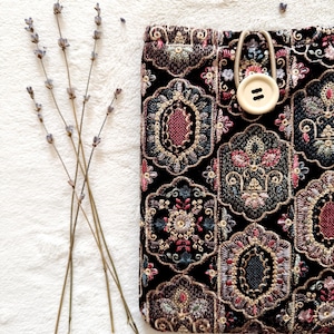 May include: Black fabric case with an ornate floral pattern in shades of red, pink, and gold. A beige button and loop closure is visible. Dried lavender sprigs are arranged on a white, textured surface.
