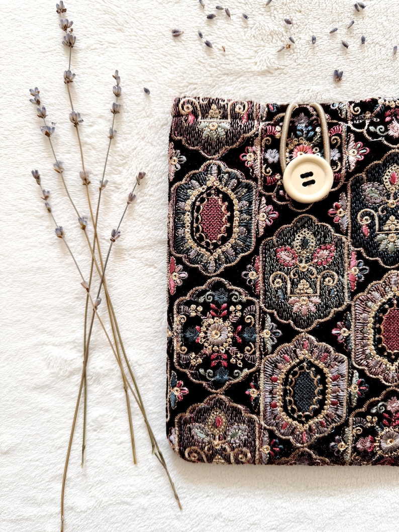 May include: Black fabric case with an ornate floral pattern in shades of red, pink, and gold. A beige button and loop closure is visible. Dried lavender sprigs are arranged on a white, textured surface.