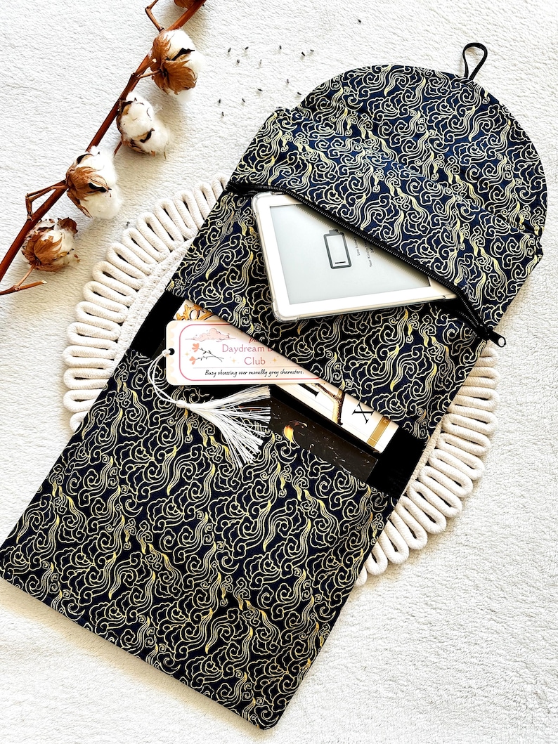 May include: A black and gold fabric pouch with a zipper closure. The pouch has a pattern of swirling clouds and is designed to hold an e-reader. The text "Daydream Club" is visible on a tag attached to the pouch.