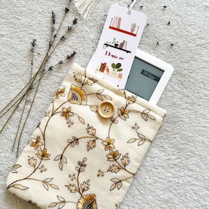 May include: A white fabric e-reader case with a floral pattern in shades of brown and yellow. The case has a button closure and a bookmark with the text "I have no" and a picture of bookshelves.