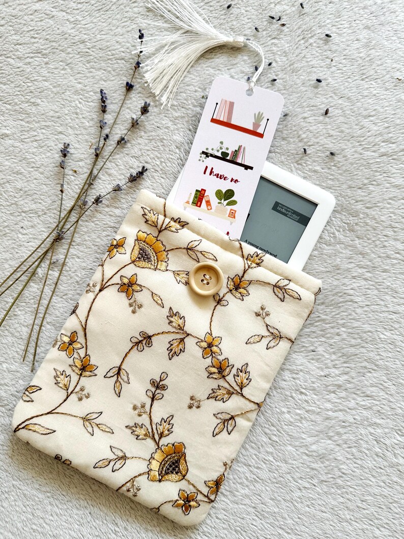 May include: A white fabric e-reader case with a floral pattern in shades of brown and yellow. The case has a button closure and a bookmark with the text "I have no" and a picture of bookshelves.