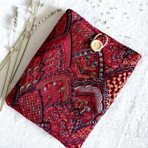 Op de afbeelding: Een bordeauxrode, geborduurde stoffen hoes met een houten knoopsluiting. De hoes heeft ingewikkelde patronen in rood, goud en zilver. Gedroogde lavendel takjes liggen naast de hoes op een witte ondergrond.