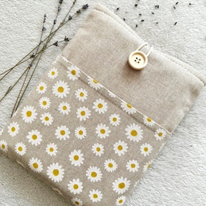 May include: A beige linen book cover with a white daisy pattern and a beige button closure. The cover is lying on a white fuzzy surface.
