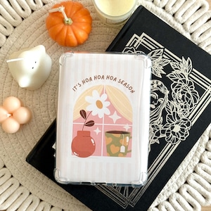 Op de afbeelding: Een doorzichtige telefoonhoes met de tekst "It's Hoa Hoa Hoa Season" over een raamontwerp met een vaas, bloem en theekopje. De hoes ligt op een zwart boek, omringd door kaarsen en een kleine pompoen.