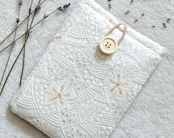 Puede incluir: Bolsa de tela blanca con un delicado patrón bordado y un cierre de botón beige. La bolsa está sobre una superficie blanca y peluda.