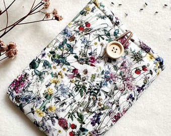Puede incluir: Funda blanca para tablet con estampado floral, con una vibrante mezcla de flores silvestres en tonos morados, amarillos, rojos y verdes. La funda se asegura con un botón de madera y una presilla elástica marrón. Textura acolchada.