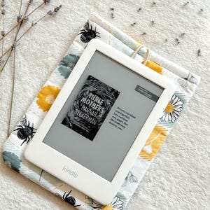 May include: A white Kindle e-reader with a floral patterned case. The screen displays the cover of the book "Three Mothers" by Hannah Beckerman. The text on the screen reads "Secrets can bind you together. Lies can tear you apart. But the truth may destroy you."