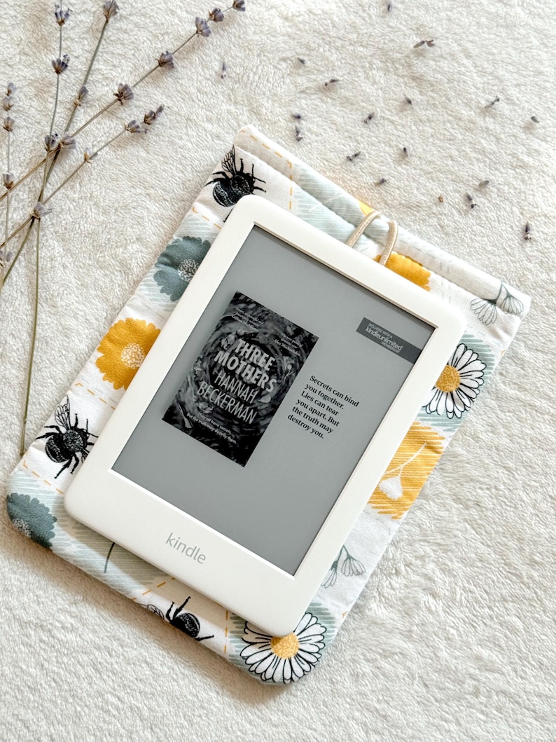 May include: A white Kindle e-reader with a floral patterned case. The screen displays the cover of the book "Three Mothers" by Hannah Beckerman. The text on the screen reads "Secrets can bind you together. Lies can tear you apart. But the truth may destroy you."