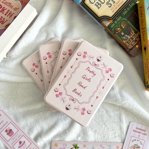 Puede incluir: Una colección de libros y marcadores sobre una superficie blanca. Los marcadores tienen varios diseños, incluyendo cupcakes, un flamenco y el texto "Coastal Girlie Essentials". El foco principal está en una pila de tarjetas rosas con el texto "Pretty Girls Read Books".