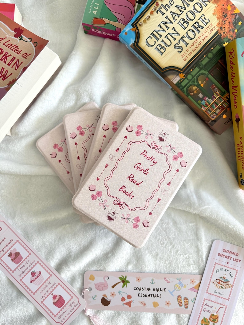 May include: A collection of books and bookmarks on a white surface. The bookmarks have various designs, including cupcakes, a flamingo, and the text "Coastal Girlie Essentials." The main focus is on a stack of pink cards with the text "Pretty Girls Read Books."
