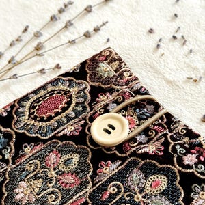 May include: A black fabric item with an ornate floral embroidery design in shades of pink, blue, and gold. A beige button with two holes and a matching elastic loop secure the item. Dried lavender sprigs are in the background.