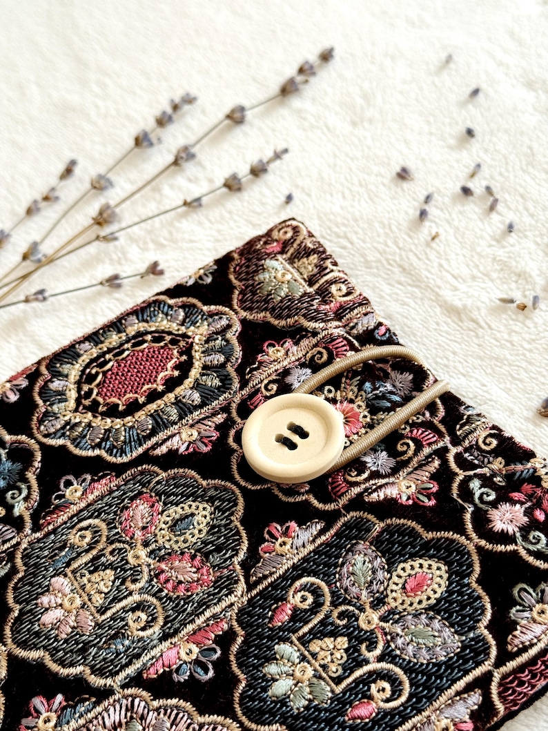 May include: A black fabric item with an ornate floral embroidery design in shades of pink, blue, and gold. A beige button with two holes and a matching elastic loop secure the item. Dried lavender sprigs are in the background.