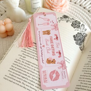 May include: A rectangular pink bookmark with a light pink tassel, featuring illustrations of two champagne glasses, two teddy bears, bows, and a lip print. The text reads "The Fake Dating Book Club", "We're just playing a role, right? No feelings involved?", and "Admit One Twisted Liar".