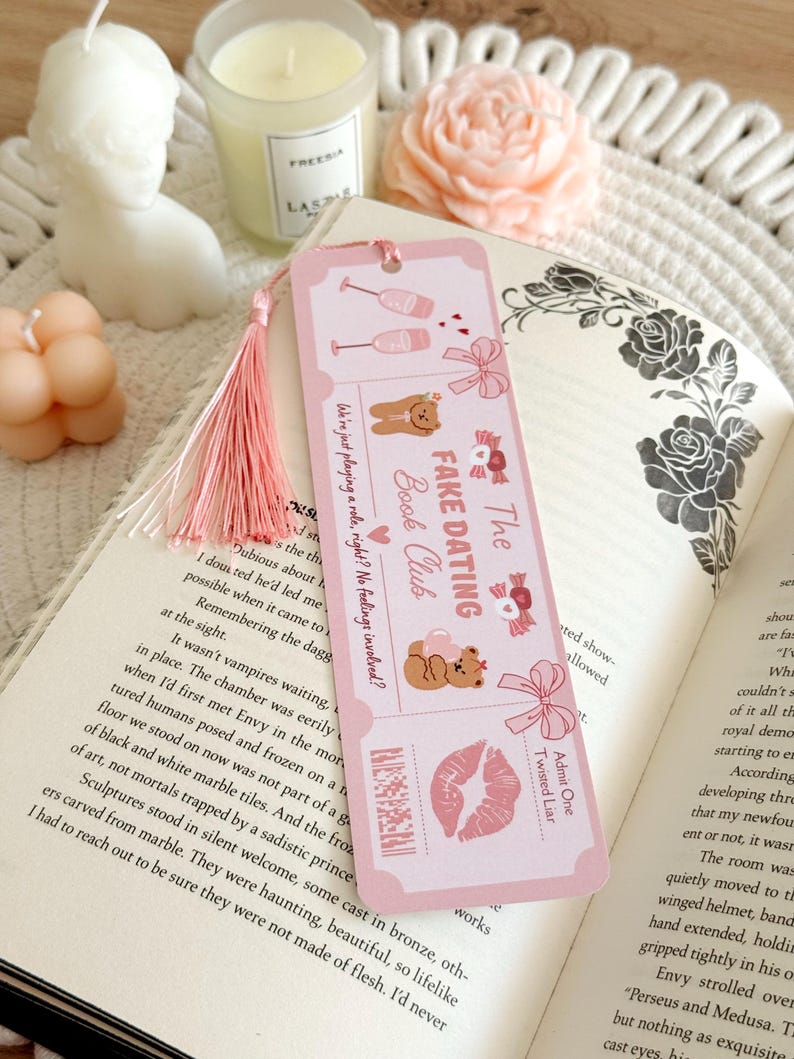 May include: A rectangular pink bookmark with a light pink tassel, featuring illustrations of two champagne glasses, two teddy bears, bows, and a lip print. The text reads "The Fake Dating Book Club", "We're just playing a role, right? No feelings involved?", and "Admit One Twisted Liar".