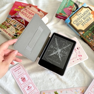 May include: An e-reader with a gray case, open to display the title "Bloodless Ties" by Katie Wismer, is surrounded by books and bookmarks. The bookmarks have cupcake and coastal themes. The books include "Love and Latters at Pumpkin Hollow" and "The Cinnamon Bun Book Store."