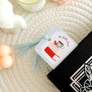 May include: A bookmark with a light blue tassel is placed in a black book. The bookmark has a graphic of books, a cup of tea, and the text "He falls first". A candle and a flower are in the background.
