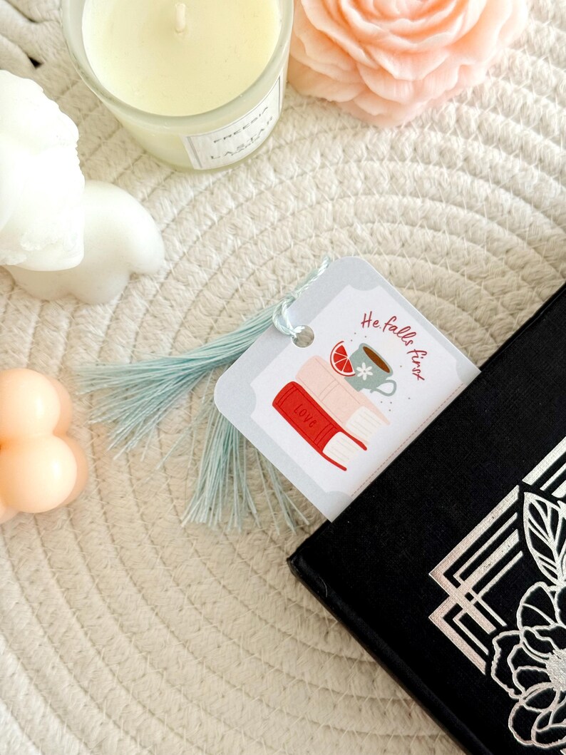 May include: A bookmark with a light blue tassel is placed in a black book. The bookmark has a graphic of books, a cup of tea, and the text "He falls first". A candle and a flower are in the background.
