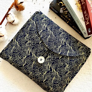 May include: A black fabric book cover with a gold cloud pattern and a white button closure. The cover is lying on a white surface.