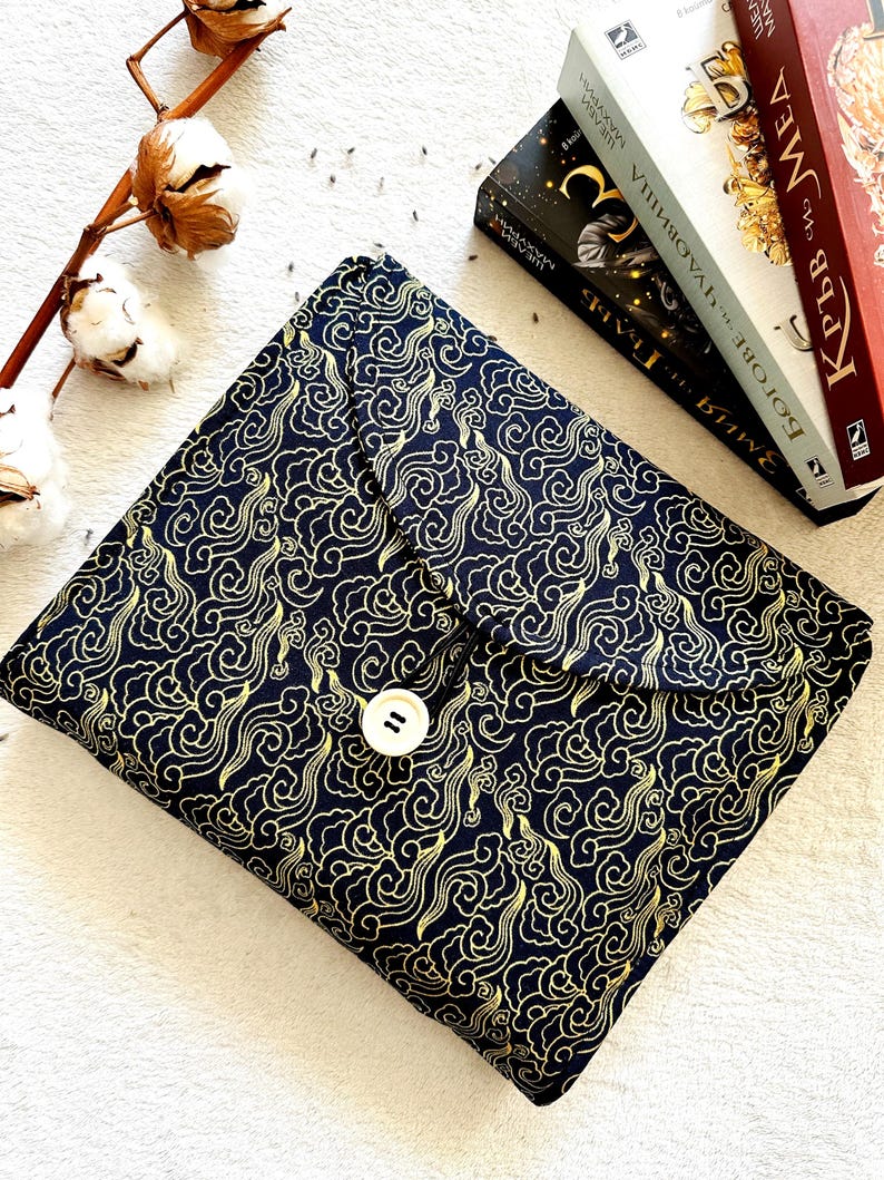 May include: A black fabric book cover with a gold cloud pattern and a white button closure. The cover is lying on a white surface.