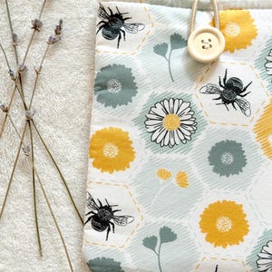 May include: A white fabric pouch with a honeycomb pattern, featuring yellow and green flowers, bees, and a button closure.