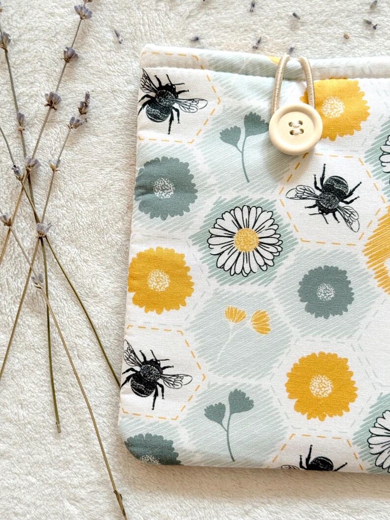 May include: A white fabric pouch with a honeycomb pattern, featuring yellow and green flowers, bees, and a button closure.