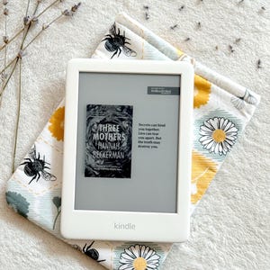 May include: A white Kindle e-reader with a gray screen displaying the cover of the book "Three Mothers" by Hannah Beckerman. The text on the screen reads "Secrets can bind you together. Lies can tear you apart. But the truth may destroy you."