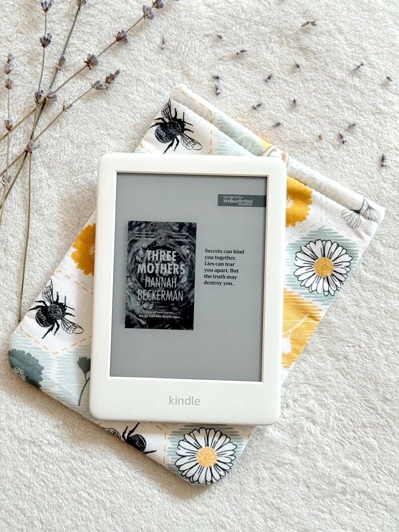 May include: A white Kindle e-reader with a gray screen displaying the cover of the book "Three Mothers" by Hannah Beckerman. The text on the screen reads "Secrets can bind you together. Lies can tear you apart. But the truth may destroy you."