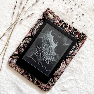 May include: A black e-reader displaying the book cover for Ruthless Ends by Katie Wismer, resting on a rectangular black fabric sleeve with intricate multi-colored floral embroidery and a small elastic loop closure.