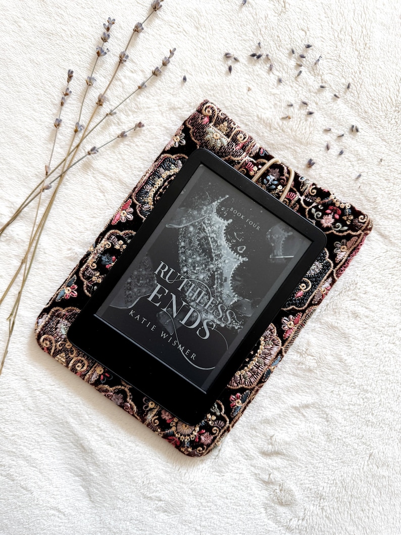 May include: A black e-reader displaying the book cover for Ruthless Ends by Katie Wismer, resting on a rectangular black fabric sleeve with intricate multi-colored floral embroidery and a small elastic loop closure.