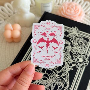 May include: A white sticker with a pink dragon design. The sticker features a dragon with wings spread, flying in the clouds. The text "THE DRAGON RIDER" is printed below the dragon. The sticker is on a black background.