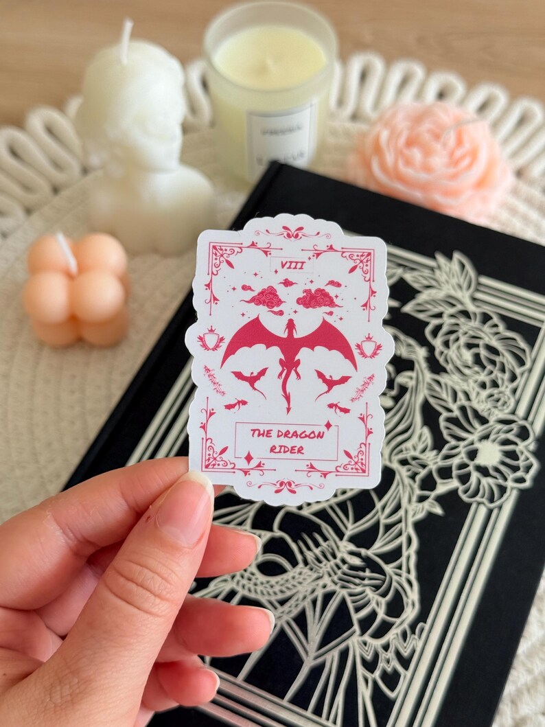 May include: A white sticker with a pink dragon design. The sticker features a dragon with wings spread, flying in the clouds. The text "THE DRAGON RIDER" is printed below the dragon. The sticker is on a black background.