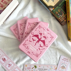 Könnte beinhalten: Ein Stapel rosa rechteckiger Schachteln mit einem Drachenmotiv und dem Text "The Dragon Rifer". Mehrere Lesezeichen mit Text und Illustrationen sind ebenfalls zu sehen, zusammen mit ein paar Büchern.