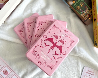 Puede incluir: Una pila de cajas rectangulares rosas con un diseño de dragón y el texto "The Dragon Rifer". También se ven varios marcadores con texto e ilustraciones, junto con algunos libros.