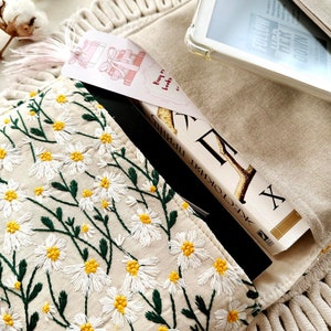 May include: A beige canvas bag with a zipper closure and a white daisy pattern embroidered on the front. A book with a black cover and a bookmark with the text "buy me books" is partially visible inside the bag. A white e-reader is also visible inside the bag.
