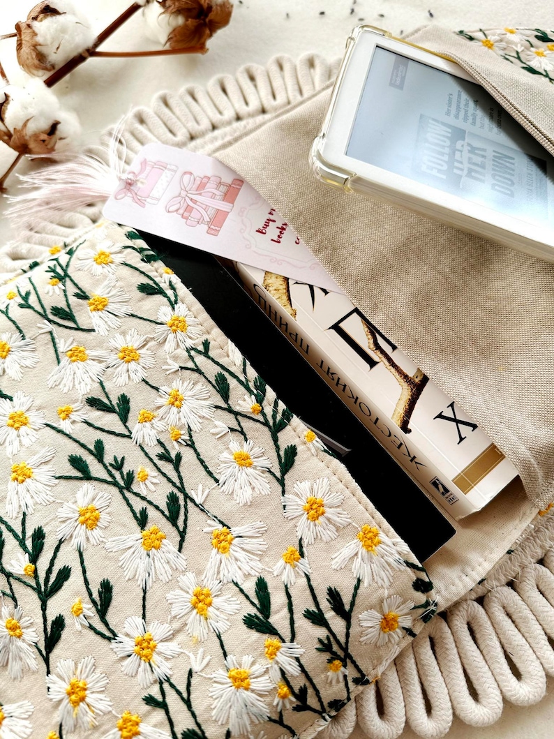May include: A beige canvas bag with a zipper closure and a white daisy pattern embroidered on the front. A book with a black cover and a bookmark with the text "buy me books" is partially visible inside the bag. A white e-reader is also visible inside the bag.