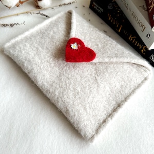 Może przedstawiać: Biała koperta z filcu z czerwonym sercem z filcu i białym kwiatem. Koperta leży na białym tle.