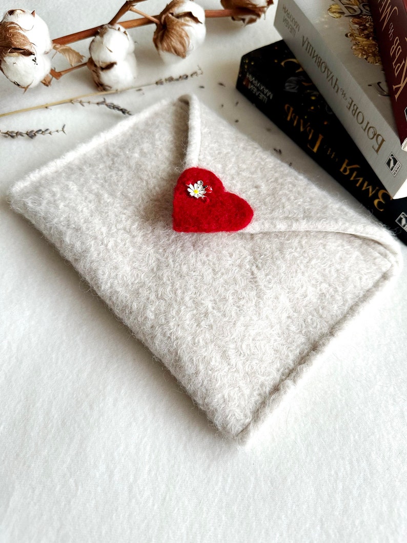 Może przedstawiać: Biała koperta z filcu z czerwonym sercem z filcu i białym kwiatem. Koperta leży na białym tle.