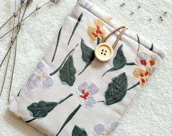 Puede incluir: Una bolsa de tela blanca con un patrón floral que presenta hojas verdes y flores moradas, amarillas y naranjas. La bolsa tiene un cierre de cordón marrón y un botón marrón.