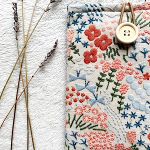 May include: A small, rectangular pouch with a floral embroidery design. The pouch is made of a natural linen fabric and features a variety of colors, including pink, orange, blue, and green. A wooden button is attached to the pouch with a brown cord.