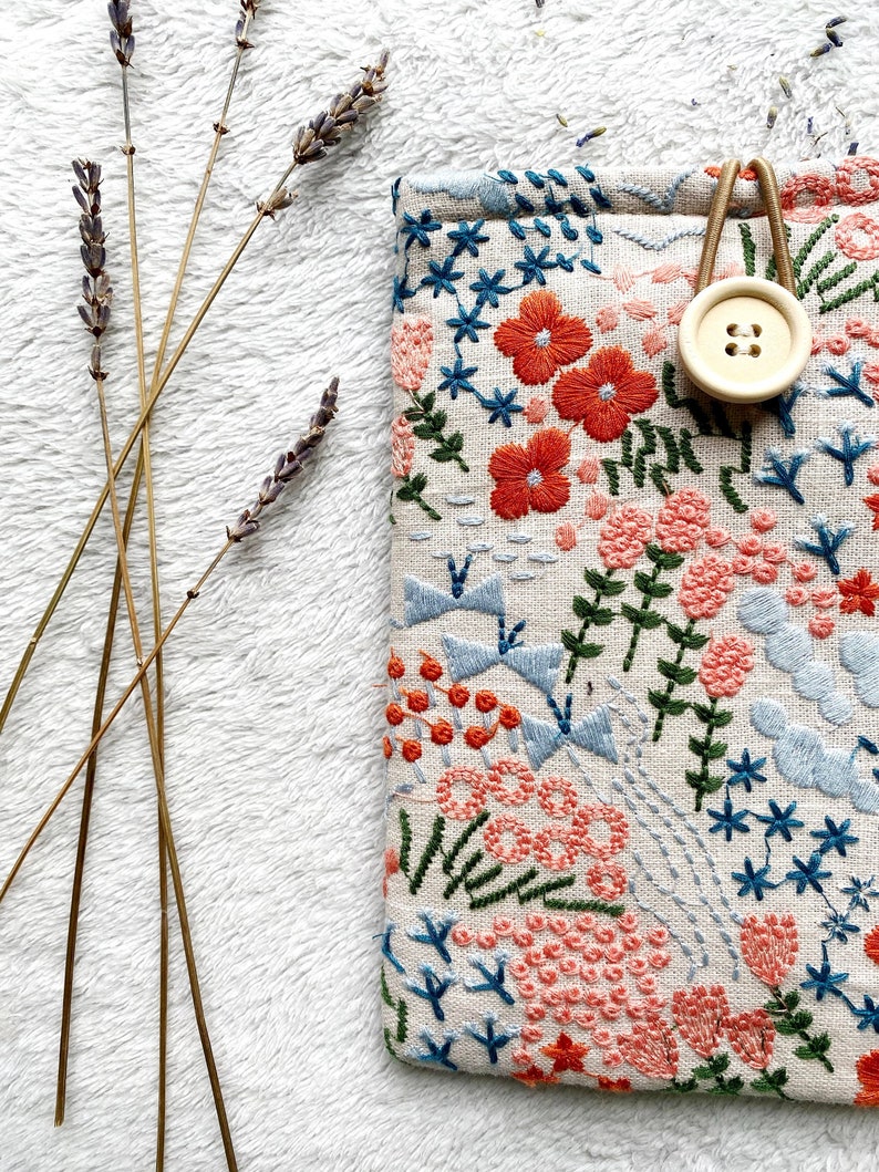 May include: A small, rectangular pouch with a floral embroidery design. The pouch is made of a natural linen fabric and features a variety of colors, including pink, orange, blue, and green. A wooden button is attached to the pouch with a brown cord.