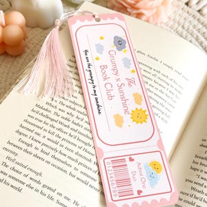 Puede incluir: Un marcap&aacute;ginas rectangular de papel con borde rosa y una borla de color rosa claro. El dise&ntilde;o presenta un gr&aacute;fico estilo entrada con el texto "The Grumpy x Sunshine Book Club" y "You are the grumpy to my sunshine," junto con ilustraciones de un sol y una nube de tormenta.