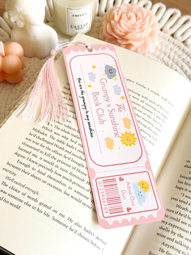 Puede incluir: Un marcap&aacute;ginas rectangular de papel con borde rosa y una borla de color rosa claro. El dise&ntilde;o presenta un gr&aacute;fico estilo entrada con el texto "The Grumpy x Sunshine Book Club" y "You are the grumpy to my sunshine," junto con ilustraciones de un sol y una nube de tormenta.